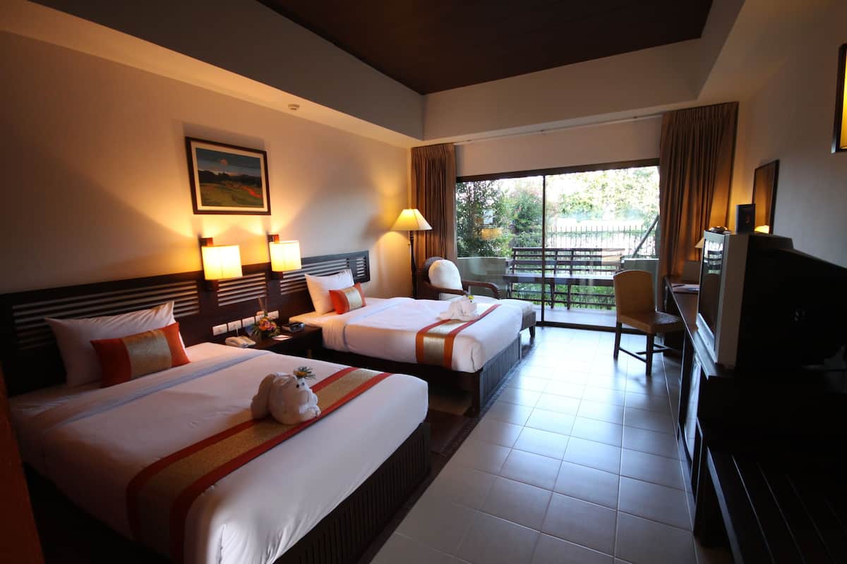 A hotel room with two beds, a desk, a chair, a television, and a balcony with a view of greenery.