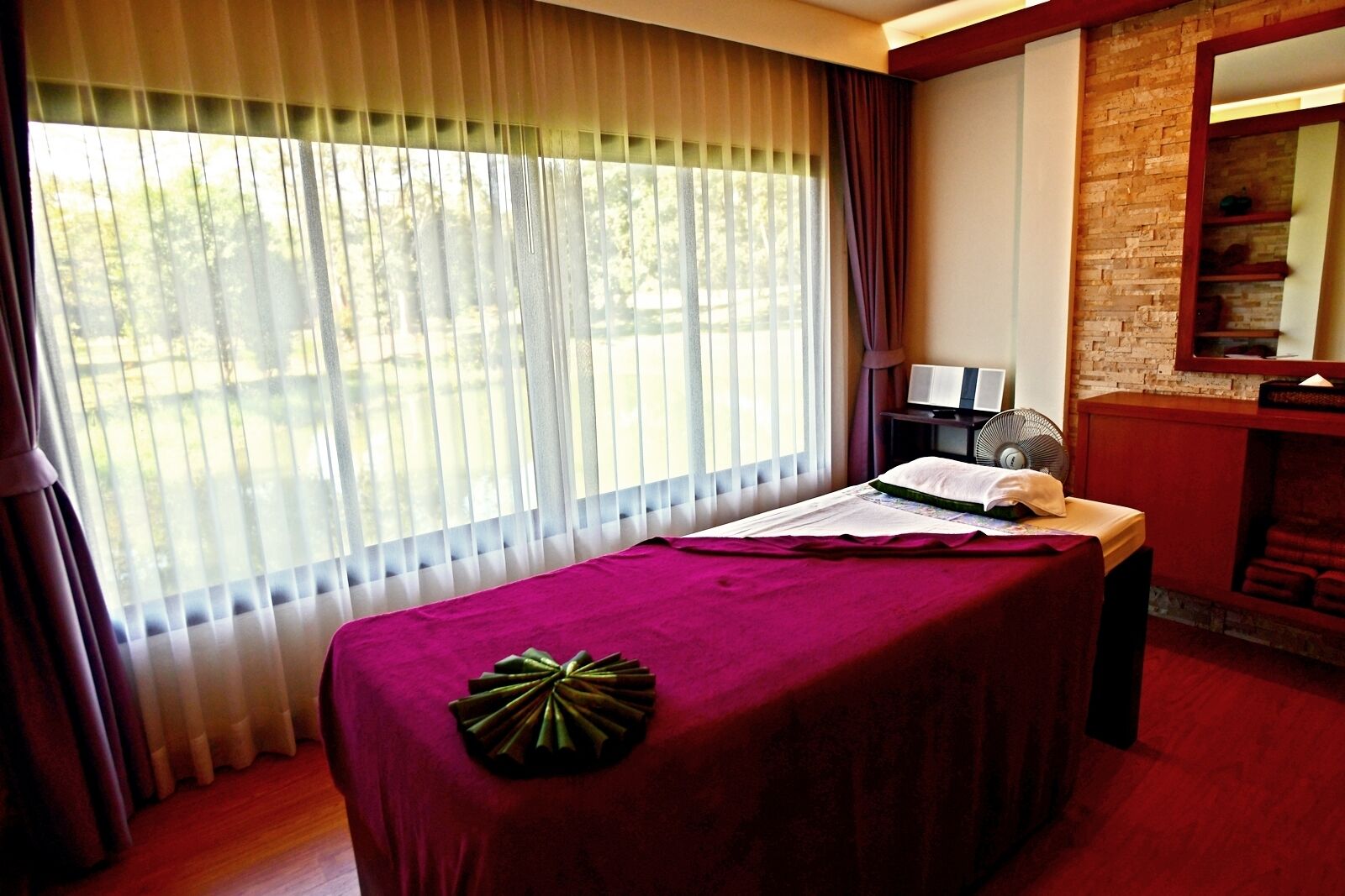 A massage table with a purple cover, a fan, a laptop, and a mirror on a brick wall.