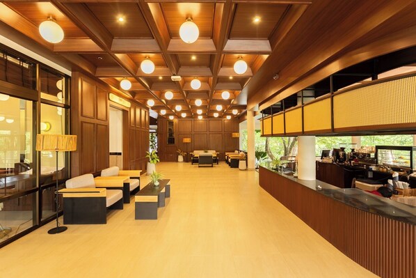 Un moderno vestíbulo de hotel con paneles de madera, luces de techo circulares, zonas de estar y una recepción con vistas a zonas verdes exteriores.