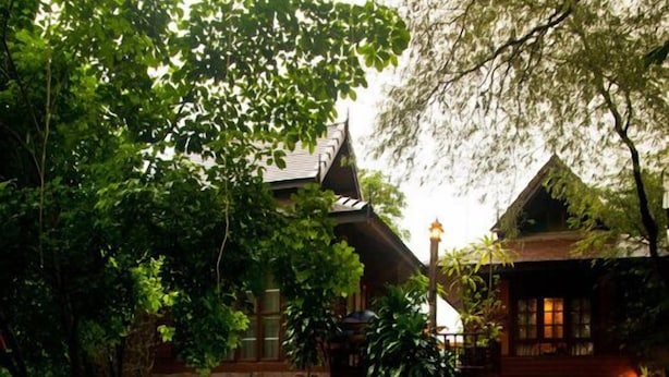 Um jardim com um elemento central de água, cercado por vegetação exuberante e um edifício tradicional com telhado inclinado.