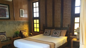Uma cama de madeira com colchão branco e almofadas estampadas, um criado-mudo com abajur e uma cadeira com almofada florida.