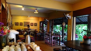 Um restaurante com área de bar, mesas e cadeiras de madeira, e um balcão com chávenas de café e uma chaleira.