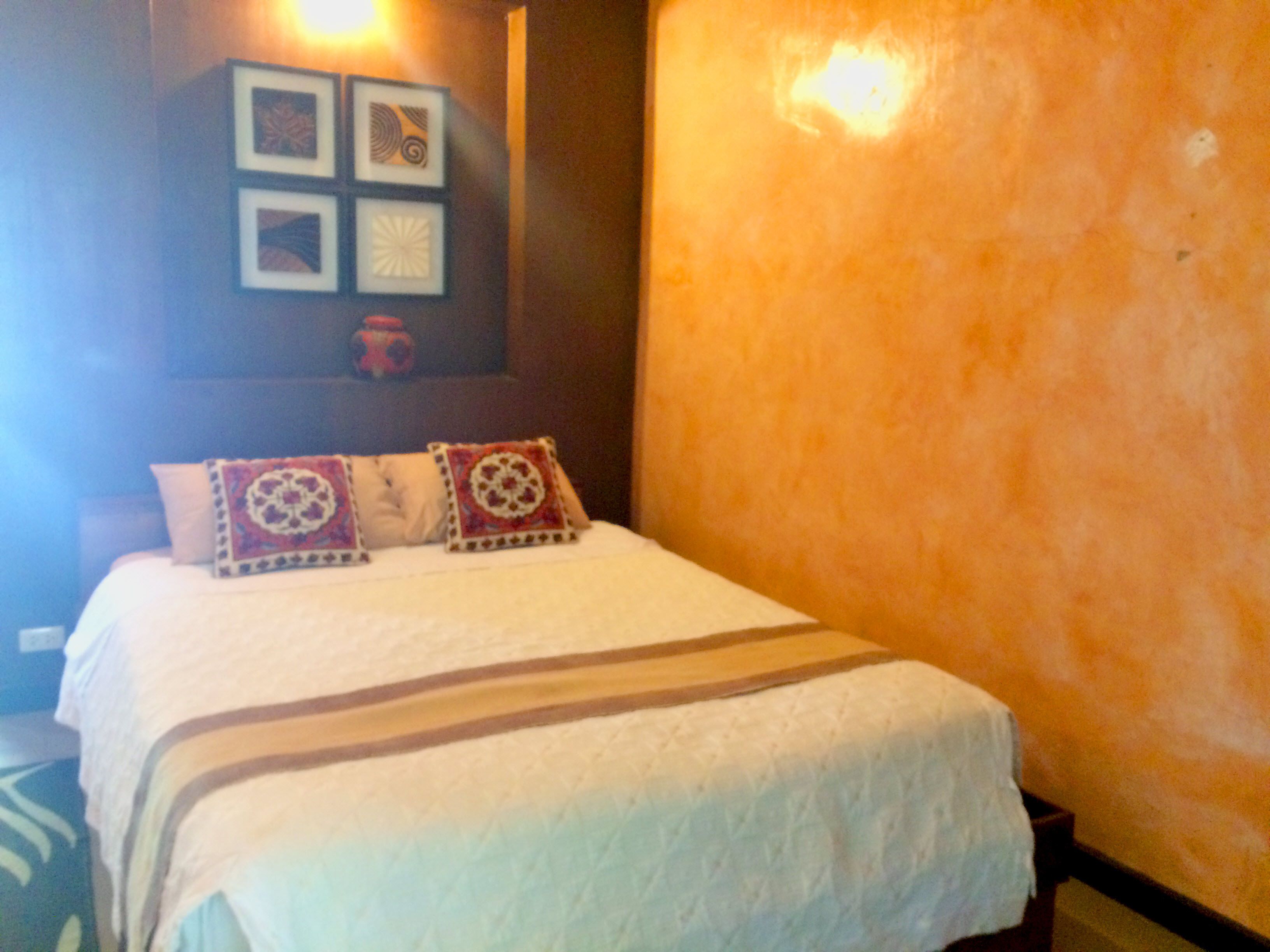 Un lit bien fait, agrémenté d’oreillers à motifs, un mur orné de tableaux encadrés et un mur orange à la texture intéressante.