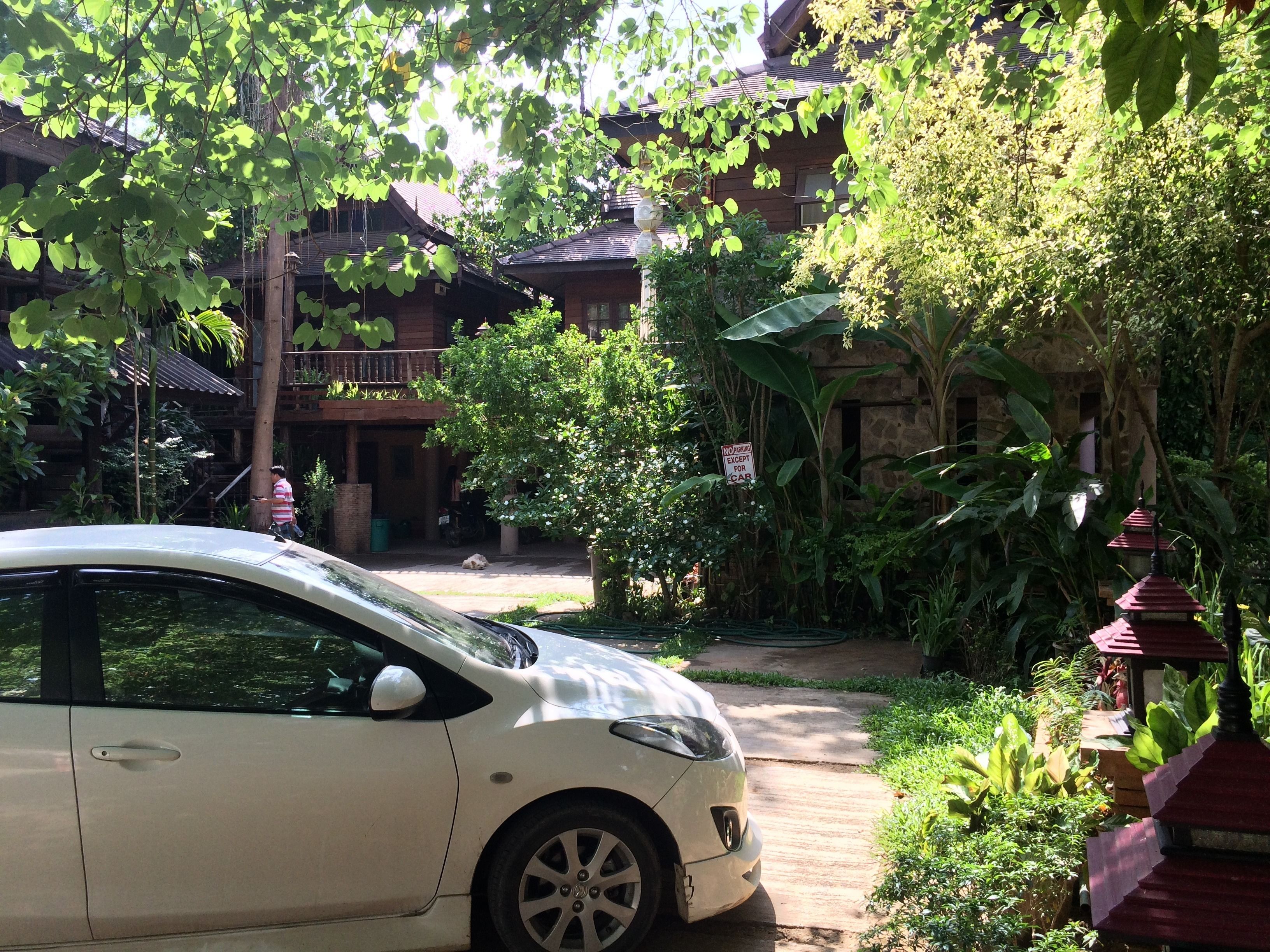 A white car parked in a lush garden area with a traditional wooden building in the background.