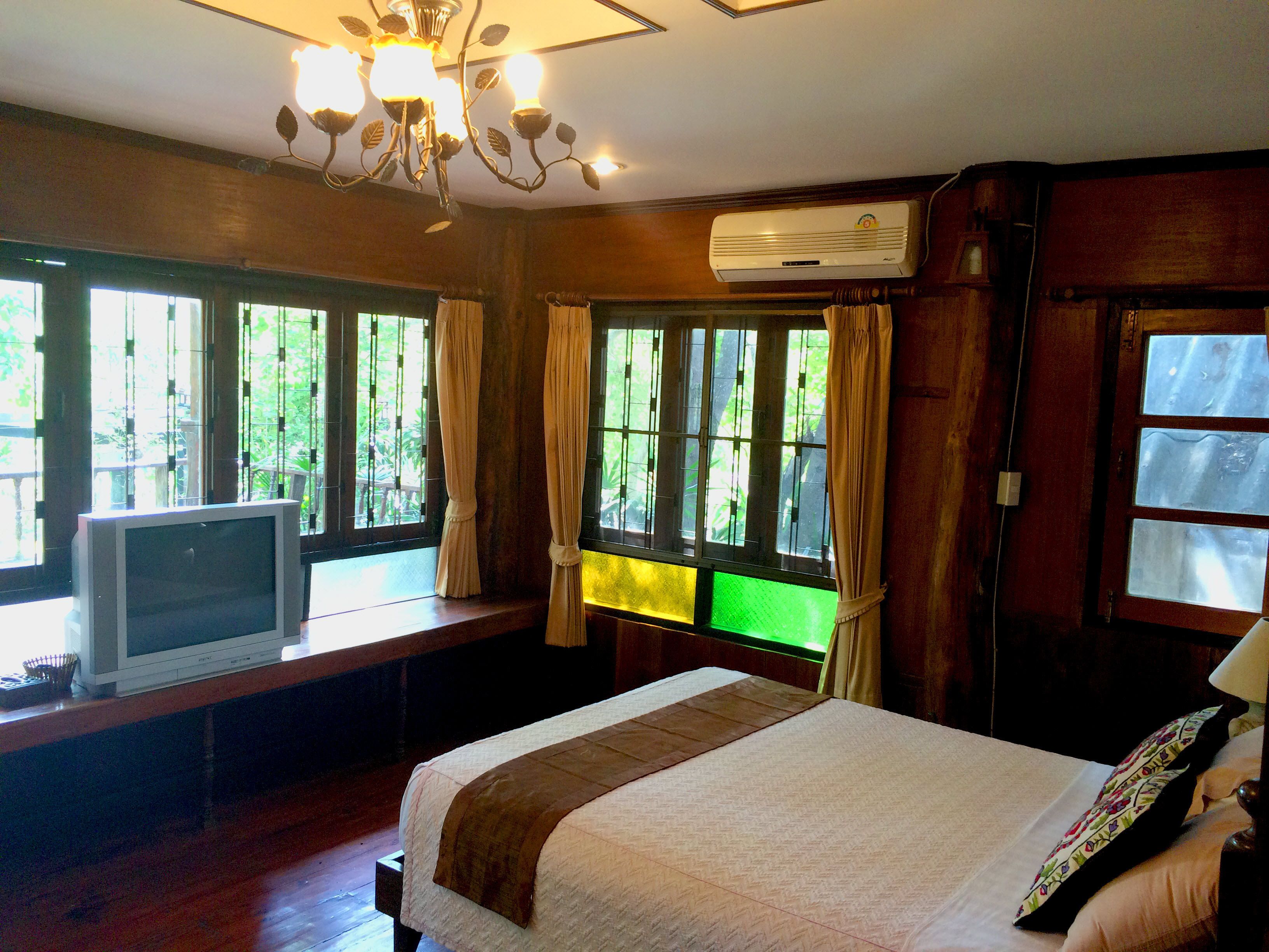 Habitación de hotel con cama, televisor sobre un banco de madera, ventanas amplias con cortinas y equipo de aire acondicionado en la pared.