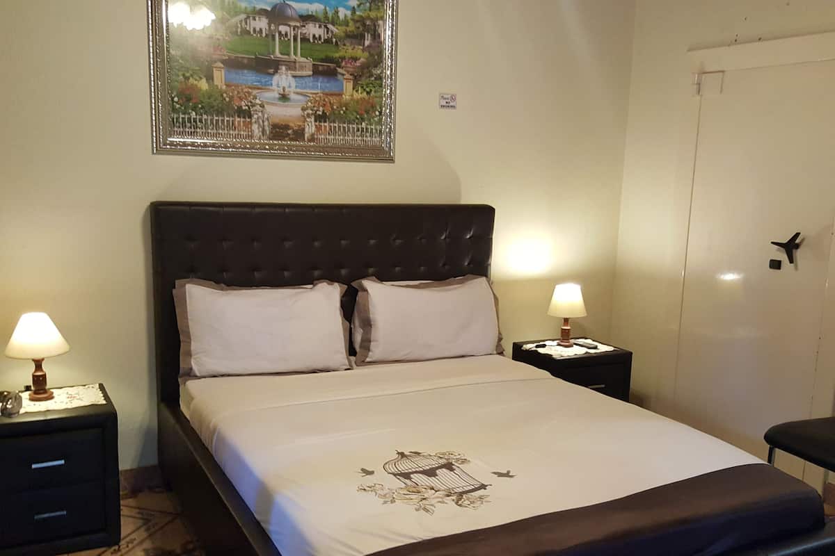 Una cama bien hecha con cabecera, dos mesitas de noche con lámparas, un cuadro enmarcado en la pared y una puerta con cámara de seguridad.