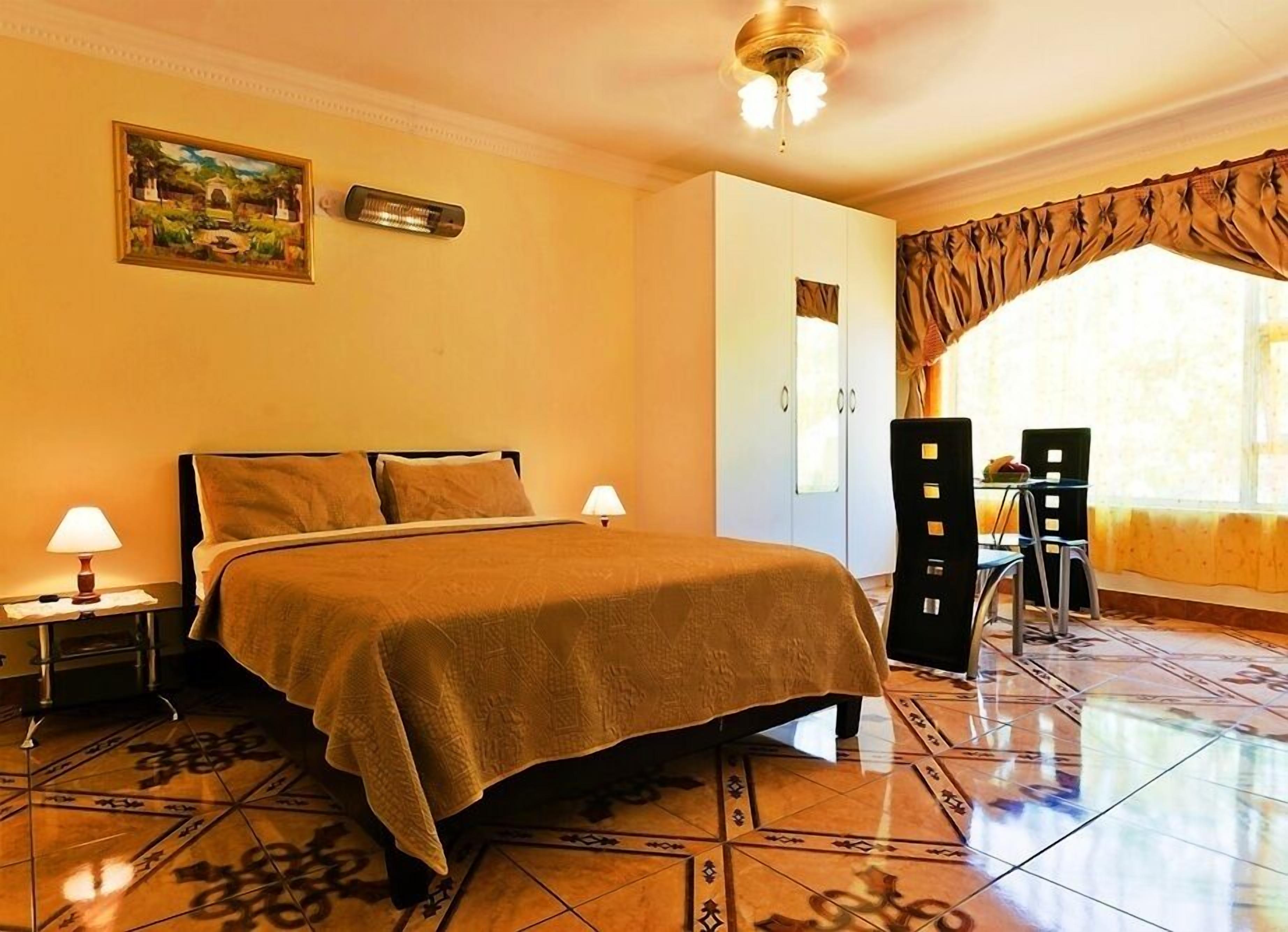 A hotel room with a bed, two bedside lamps, a painting on the wall, a wardrobe, and a dining area with chairs and a table.