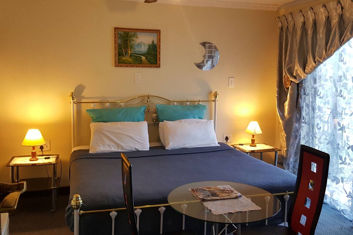 Habitación de hotel con cama, dos lámparas de noche, ventilador de techo, un cuadro, una mesa redonda con una revista y una ventana con cortinas.