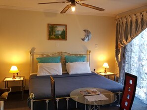 A hotel room with a bed, two bedside lamps, a ceiling fan, a painting, a round table with a magazine, and a window with curtains.