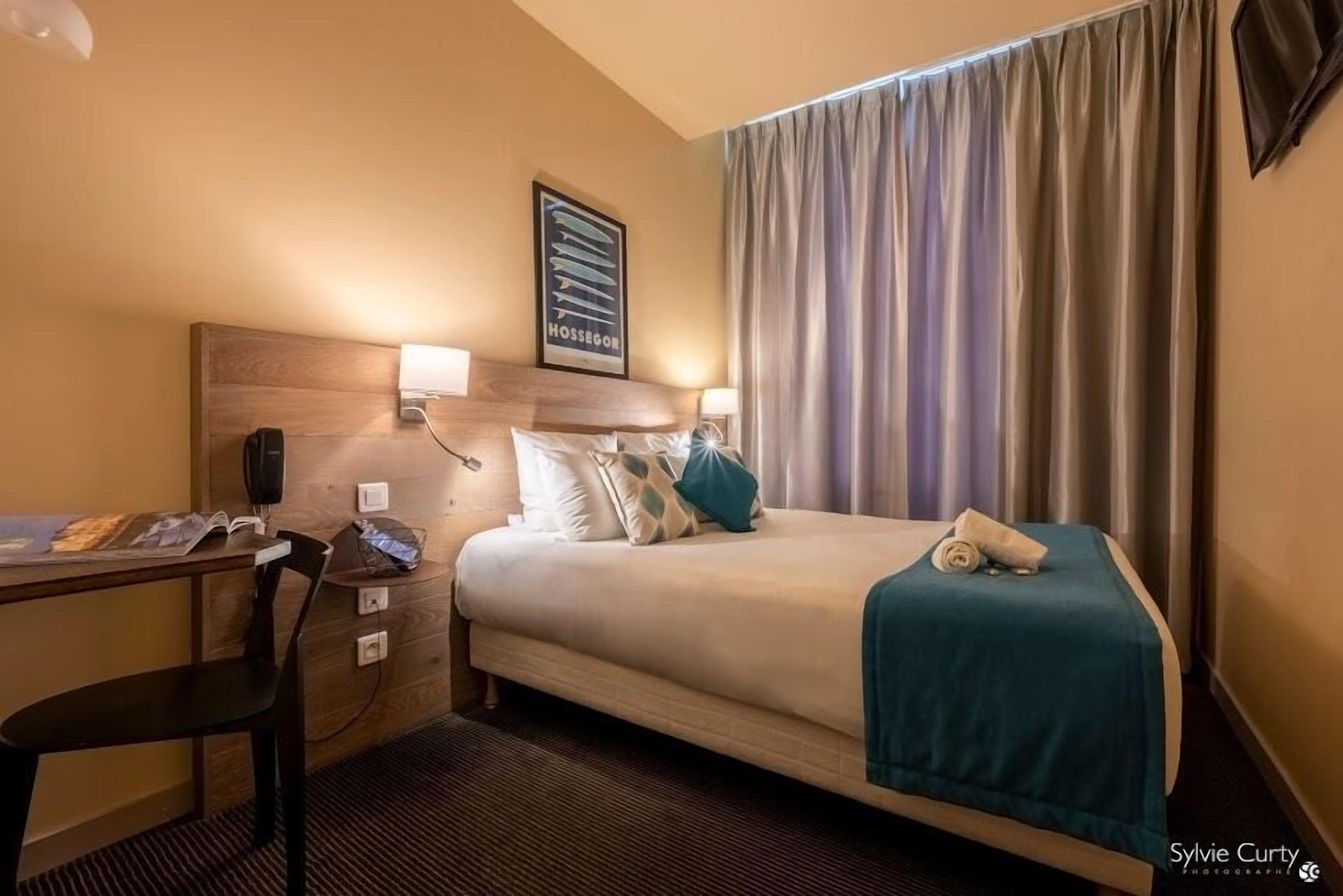 A hotel room with a bed, a desk, a chair, a lamp, and a curtain.