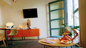 A modern living room with a flat-screen TV, a wooden coffee table, a patterned sofa, a dining area with a chair, and a cabinet. - Accostage Hotel (La Rochelle)