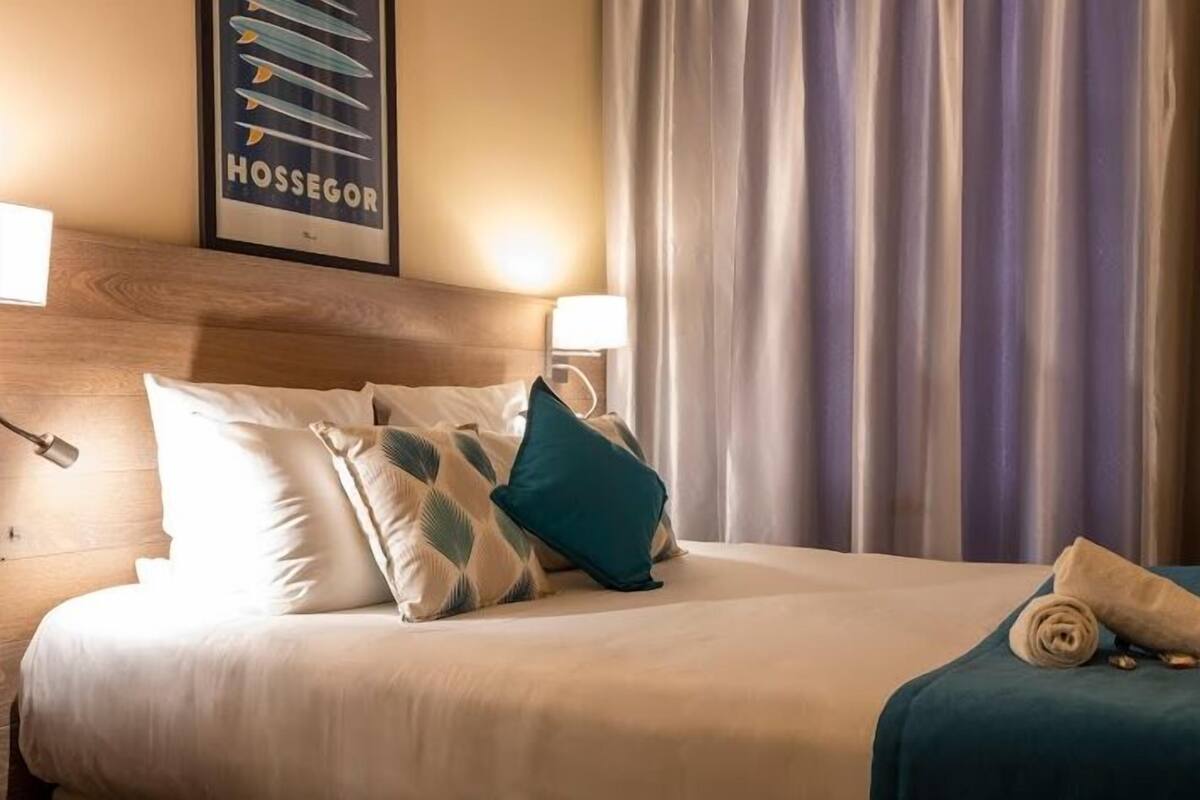 A hotel room with a neatly made bed, a headboard, a framed picture on the wall, and a window with curtains.
