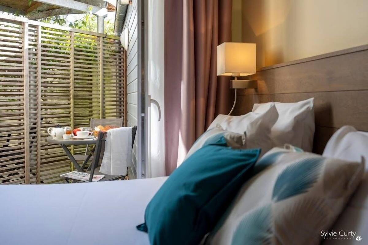 A hotel room with a bed, a table with a teapot and cups, and a view of a balcony with a wooden fence.