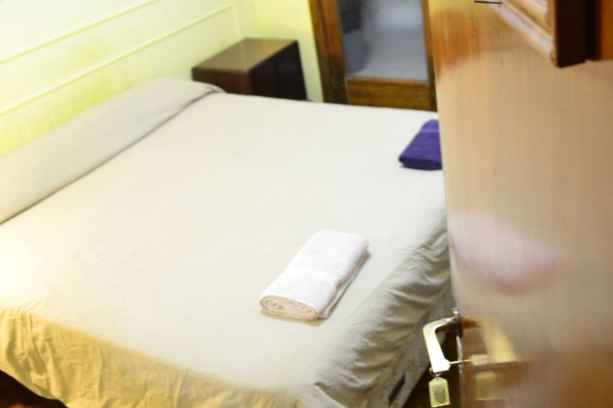 A hotel room with a bed, a wooden nightstand, a door with a frosted glass panel, and a framed picture on the wall.