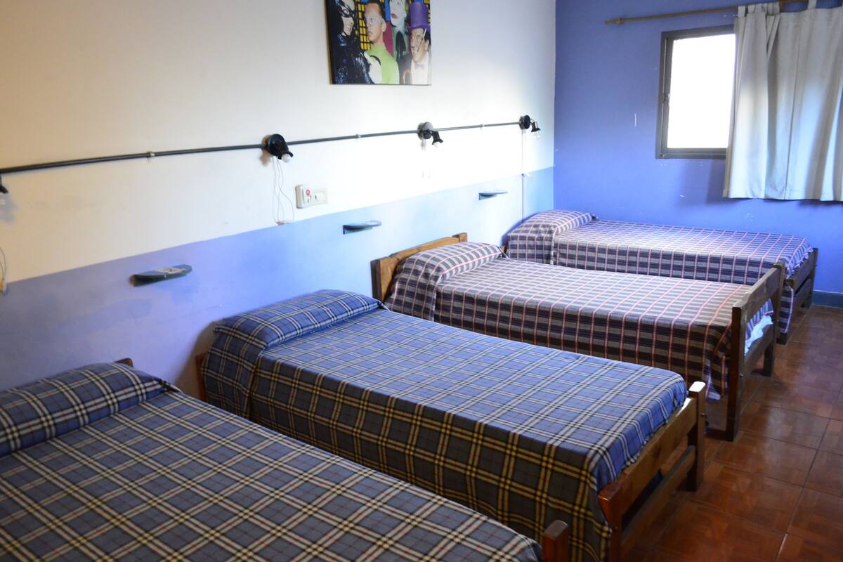 A room with three beds, each with a plaid bedspread, a window with curtains, and a wall-mounted light fixture.