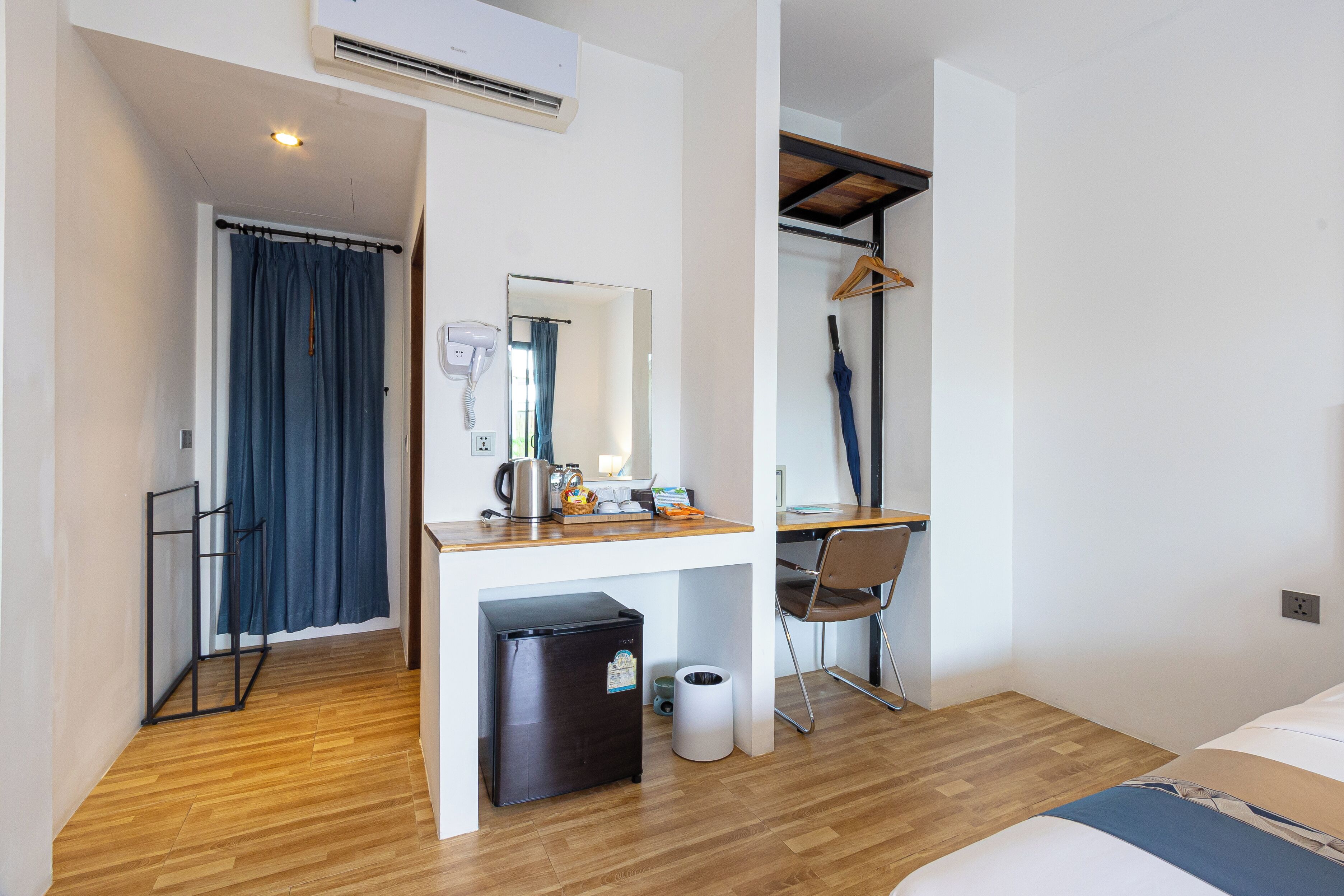 A compact hotel room with a wooden floor, a bed, a desk with a chair, a mirror, a hanging coat rack, and a small refrigerator.