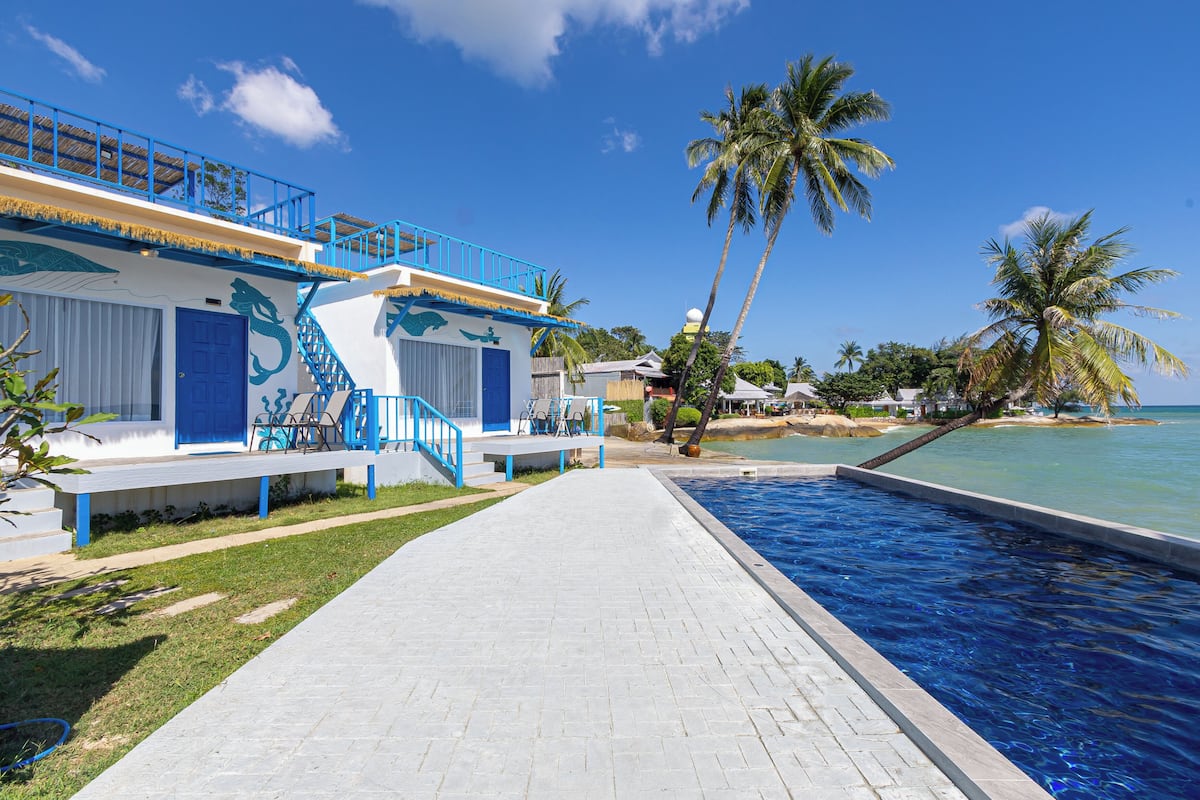 Uma área à beira da piscina com vista clara para o oceano, um edifício azul e branco com escadas e palmeiras.