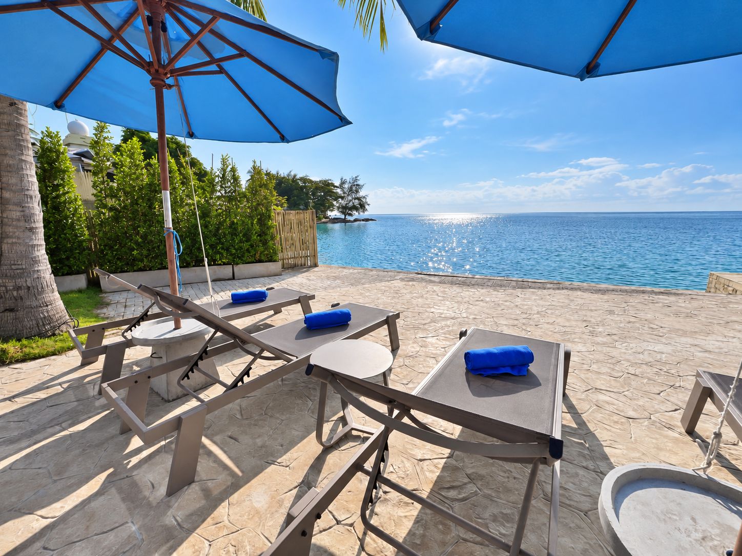 An outdoor seating area with tables and chairs, a large umbrella, and a view of the sea.