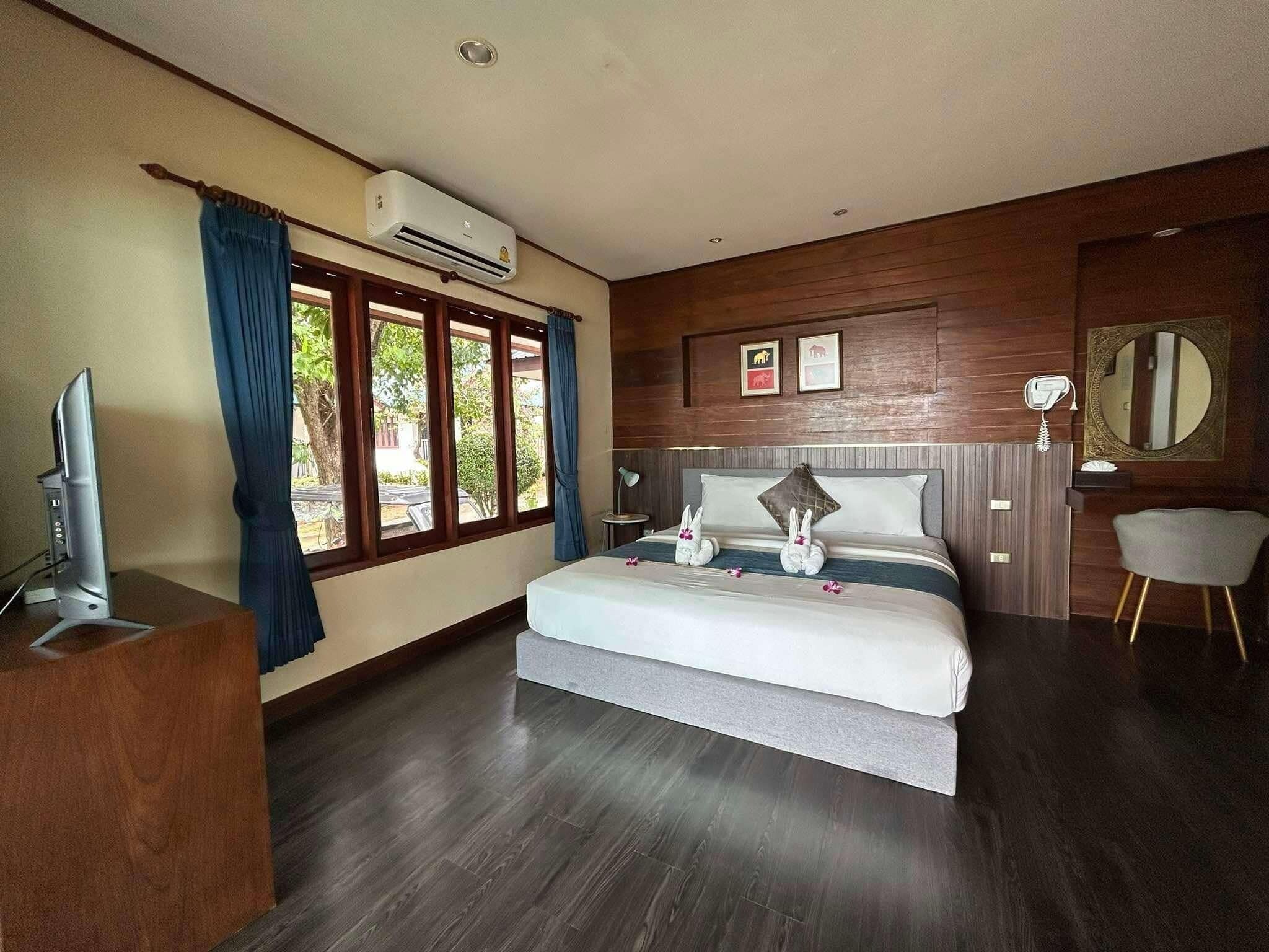 A bedroom with a bed, a TV on a wooden stand, a window with curtains, an air conditioning unit, a mirror, and a small table with a lamp.