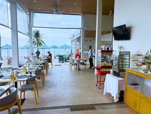 Restaurante com área externa voltada para o mar, espaço para buffet e televisão na parede.