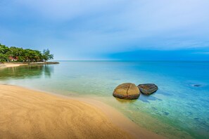 A beach with clear water, two large rocks, and a distant shoreline with buildings. - Three Trees Samui Resort - Sarocha Villa (Koh Samui)
