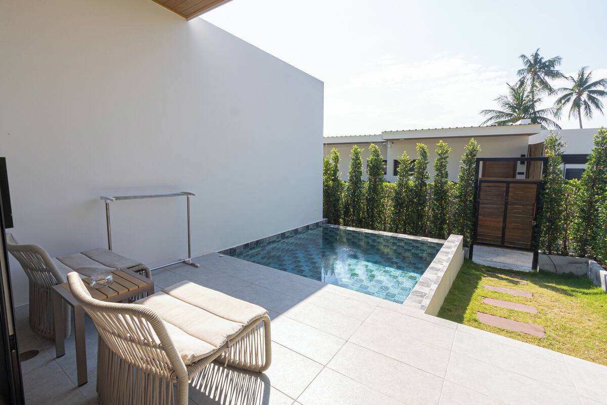 Uma área de piscina moderna ao ar livre, com espreguiçadeiras e uma pequena mesa, cercada por um muro branco e uma cerca de madeira.