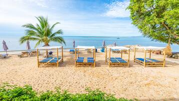 Una playa con varias cabañas de madera con cojines azules, una palmera y una vista despejada del mar.