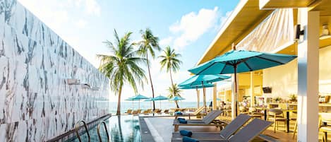 A pool area with lounge chairs and umbrellas, palm trees, and a view of the ocean.