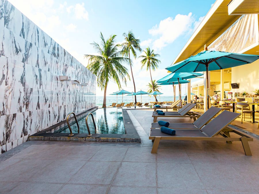 A pool area with lounge chairs and umbrellas, palm trees, and a view of the ocean.