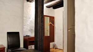 Um quarto com cama, uma cadeira, escrivaninha de madeira, espelho e porta.