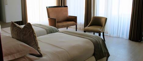 Una habitación de hotel con dos camas, una zona de estar con una silla y ventanas grandes con cortinas.
