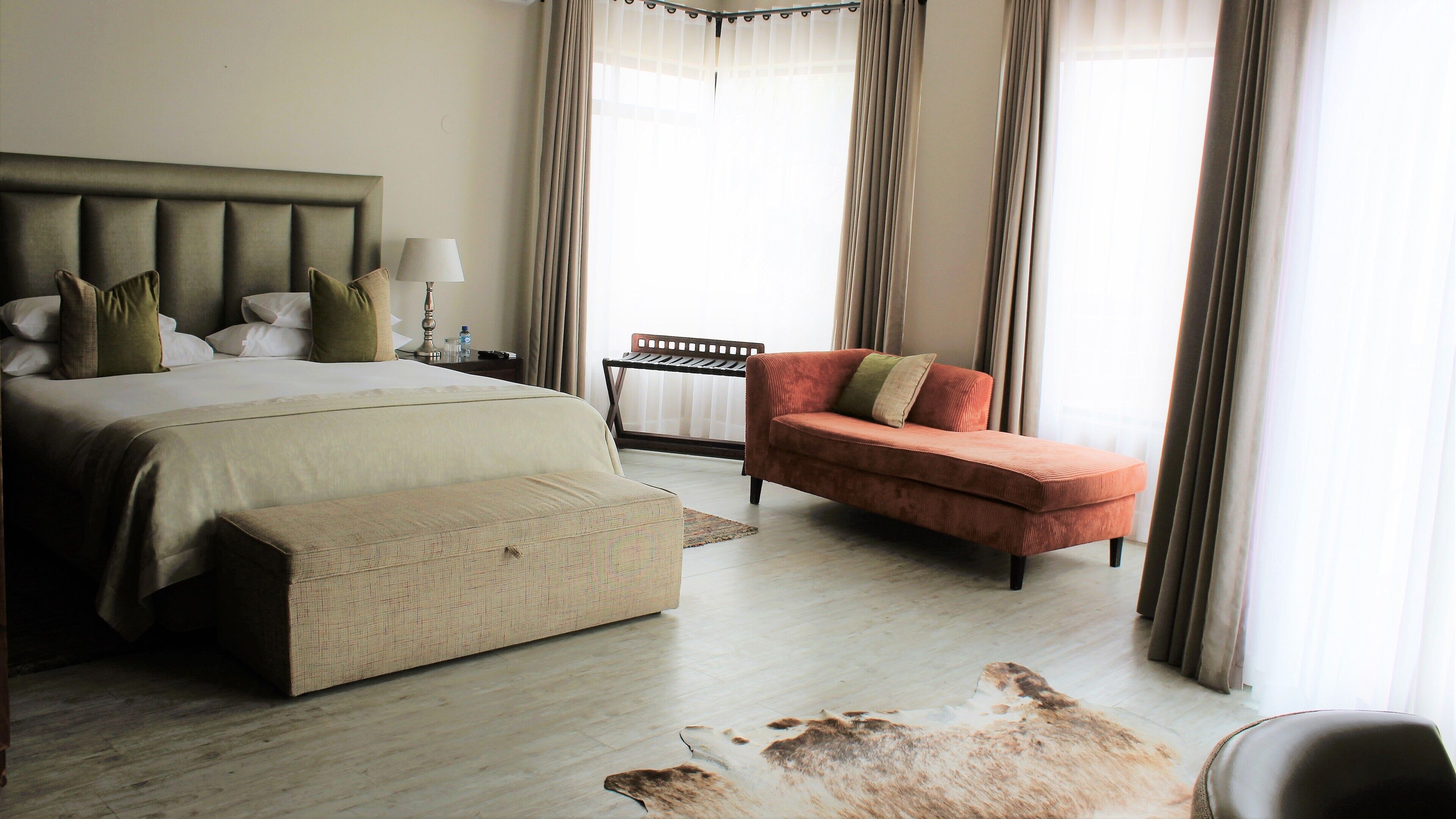 A hotel room with a bed, a red armchair, a small table with a lamp, and a window with curtains.