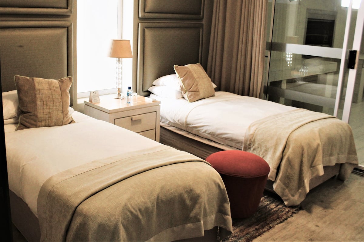 A hotel room with two beds, a nightstand, a lamp, and a red ottoman.