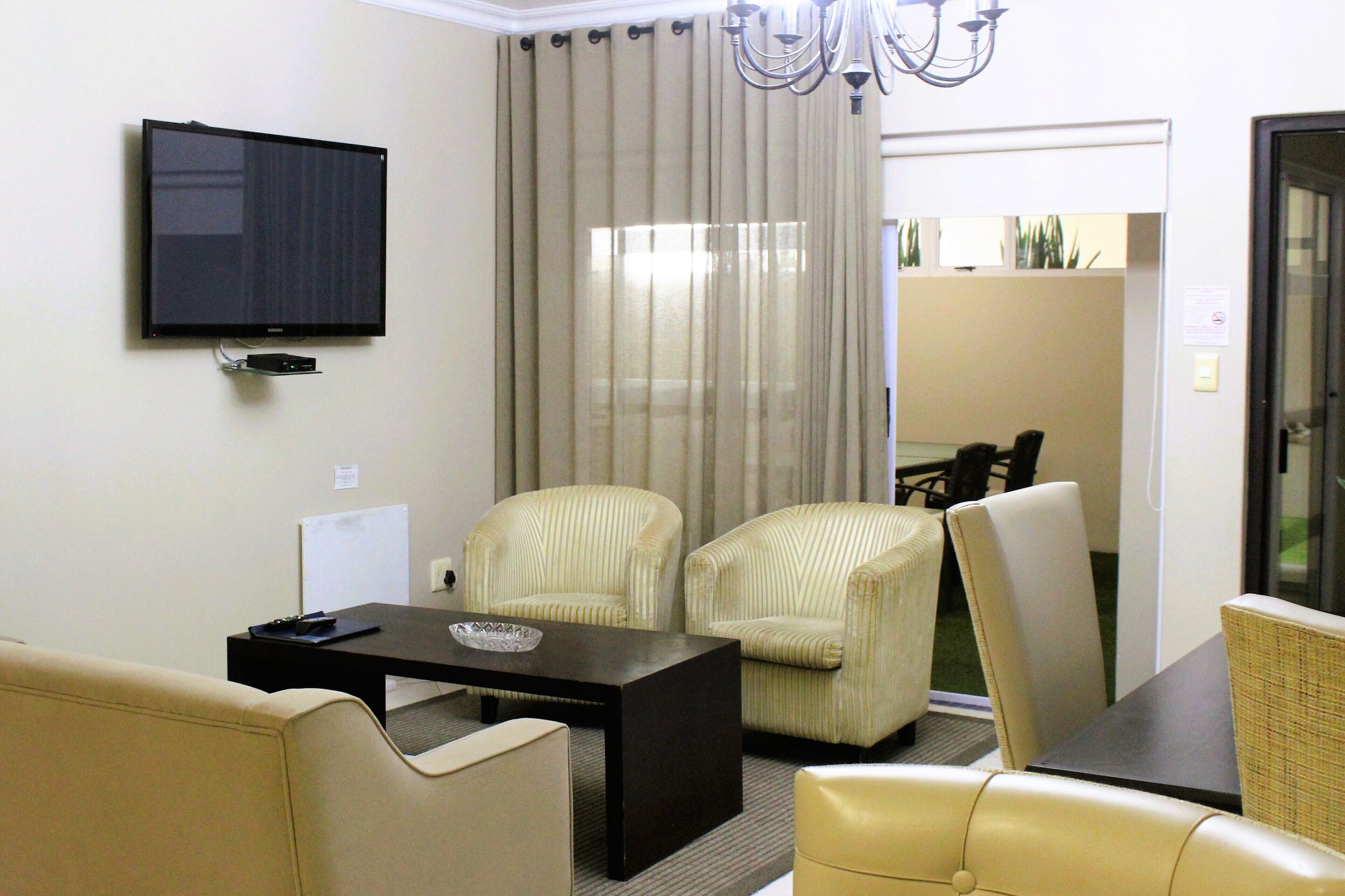 A hotel room with a flat-screen TV mounted on the wall, a dining table with chairs, a sofa, and a large window with curtains.