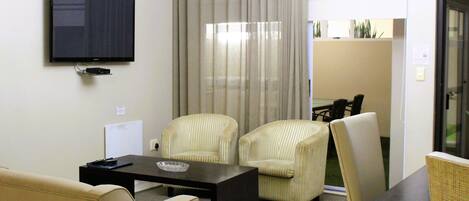 A hotel room with a flat-screen TV mounted on the wall, a dining table with chairs, a sofa, and a large window with curtains.