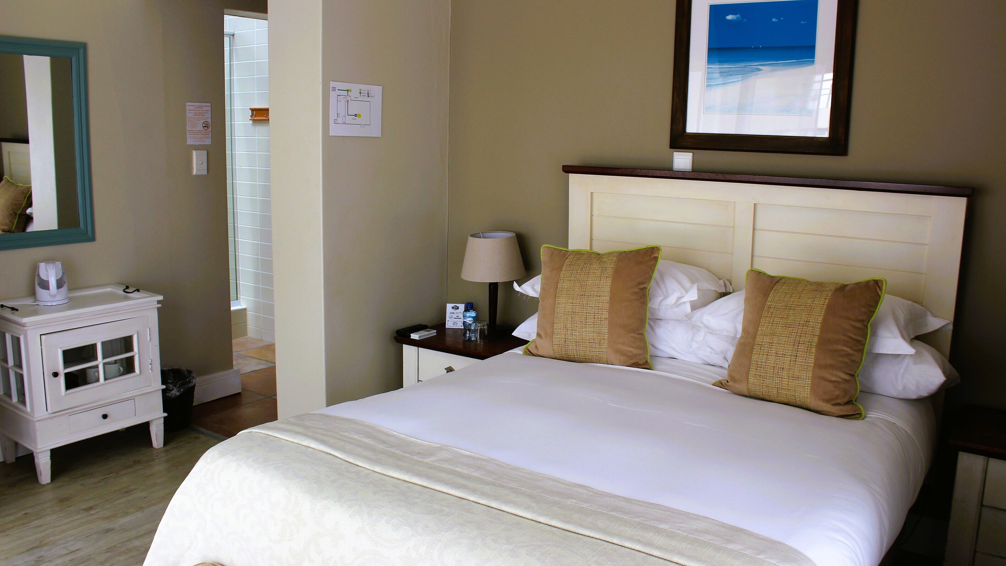 A hotel room with a bed, a nightstand with a lamp, a small table with a kettle, and a mirror on the wall.