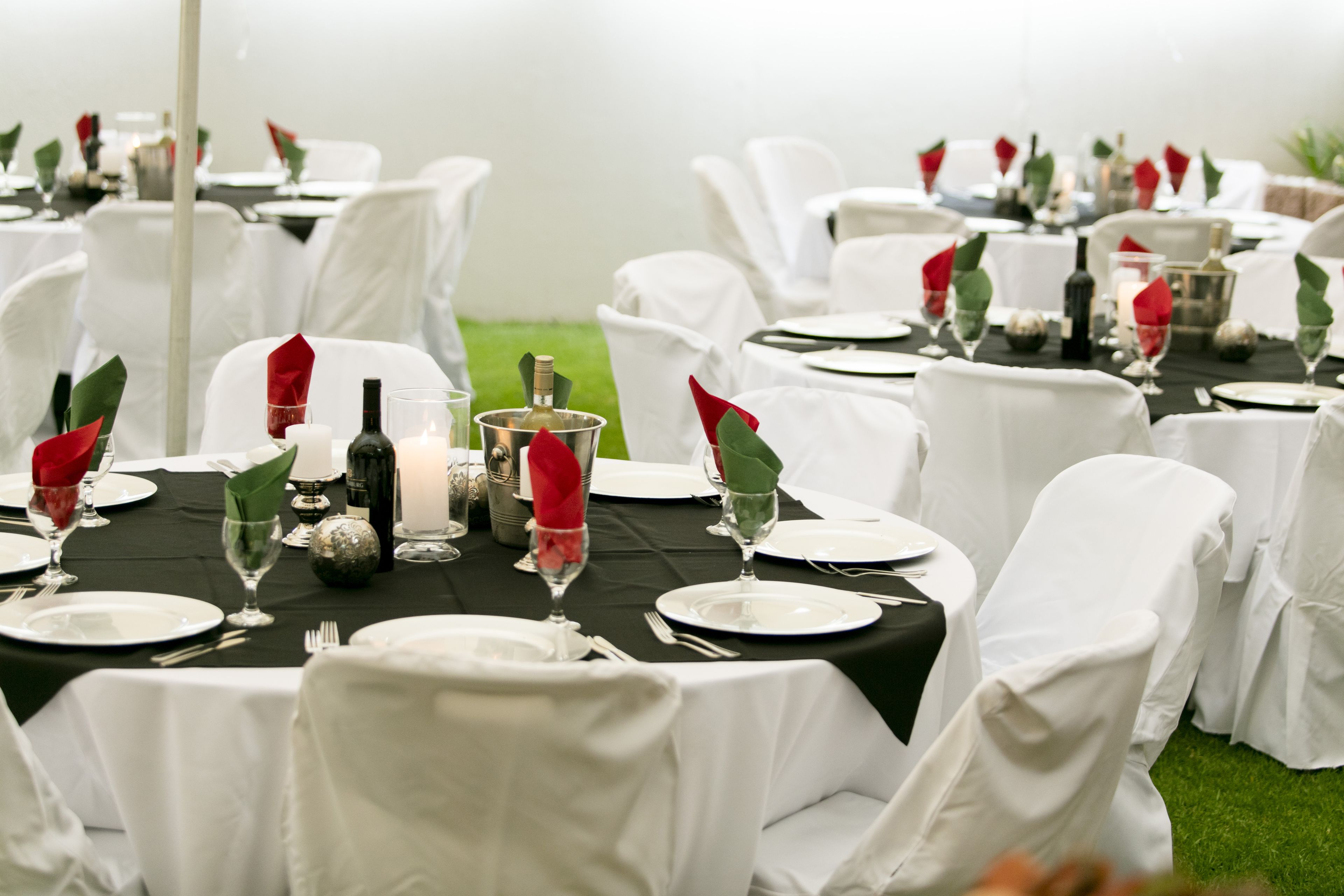 Un montaje formal para comedor al aire libre, con mesas redondas cubiertas con manteles negros, sillas blancas y servilletas rojas.