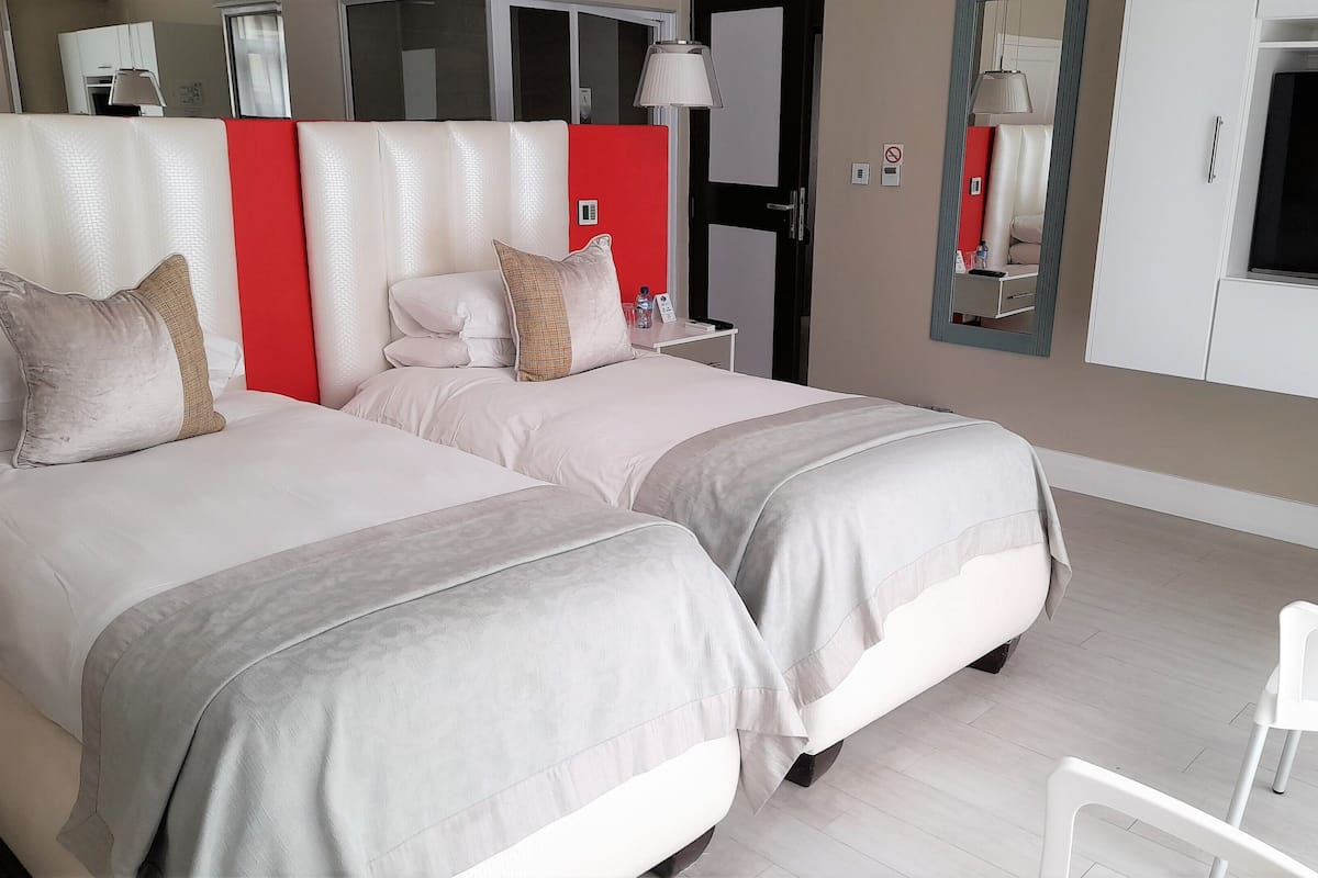 A hotel room with two beds, a red accent wall, a bedside table with a lamp, and a large mirror.