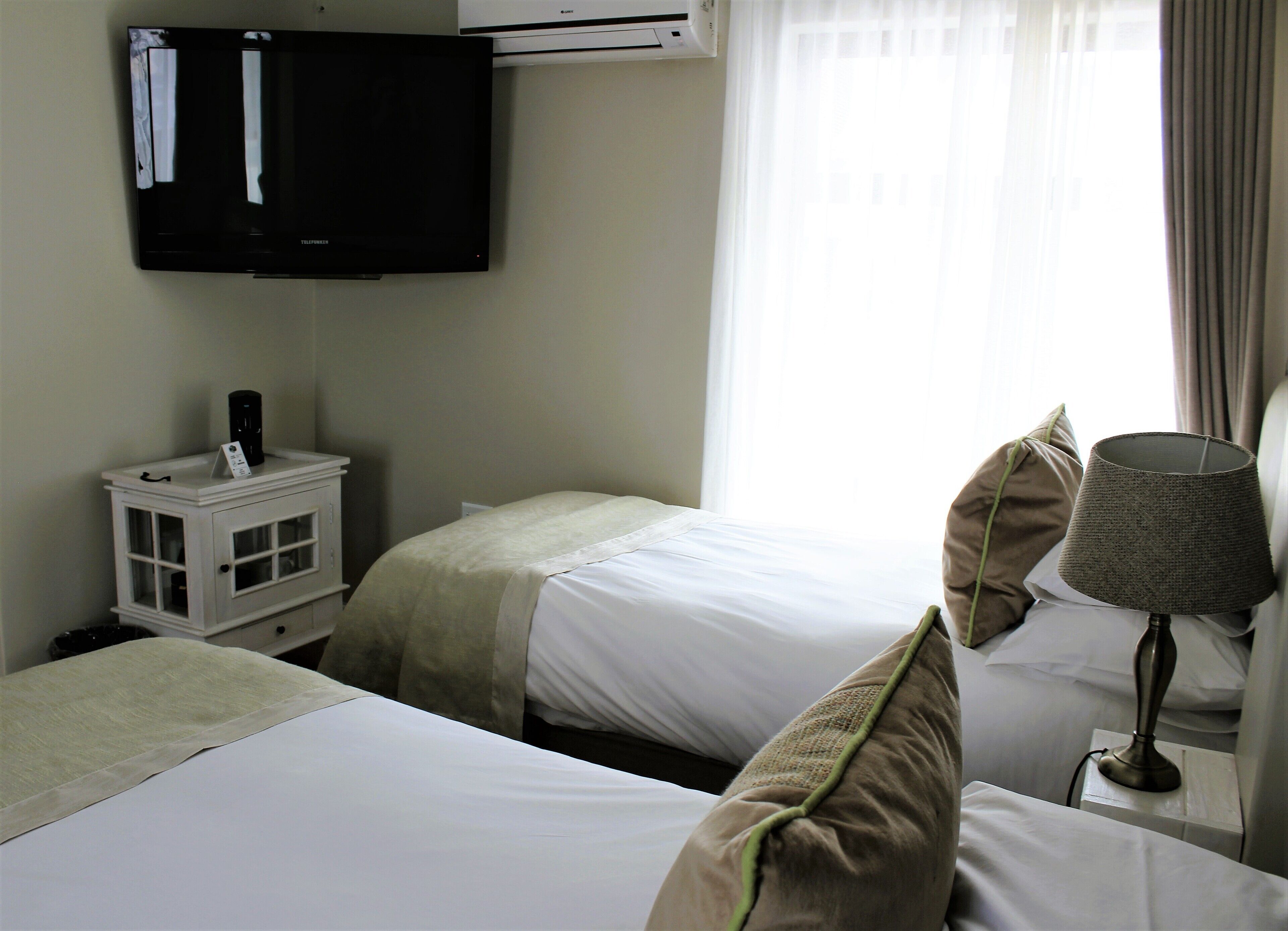 Une chambre d’hôtel avec deux lits, un téléviseur fixé au mur, une table de chevet avec une lampe et une fenêtre avec des rideaux.