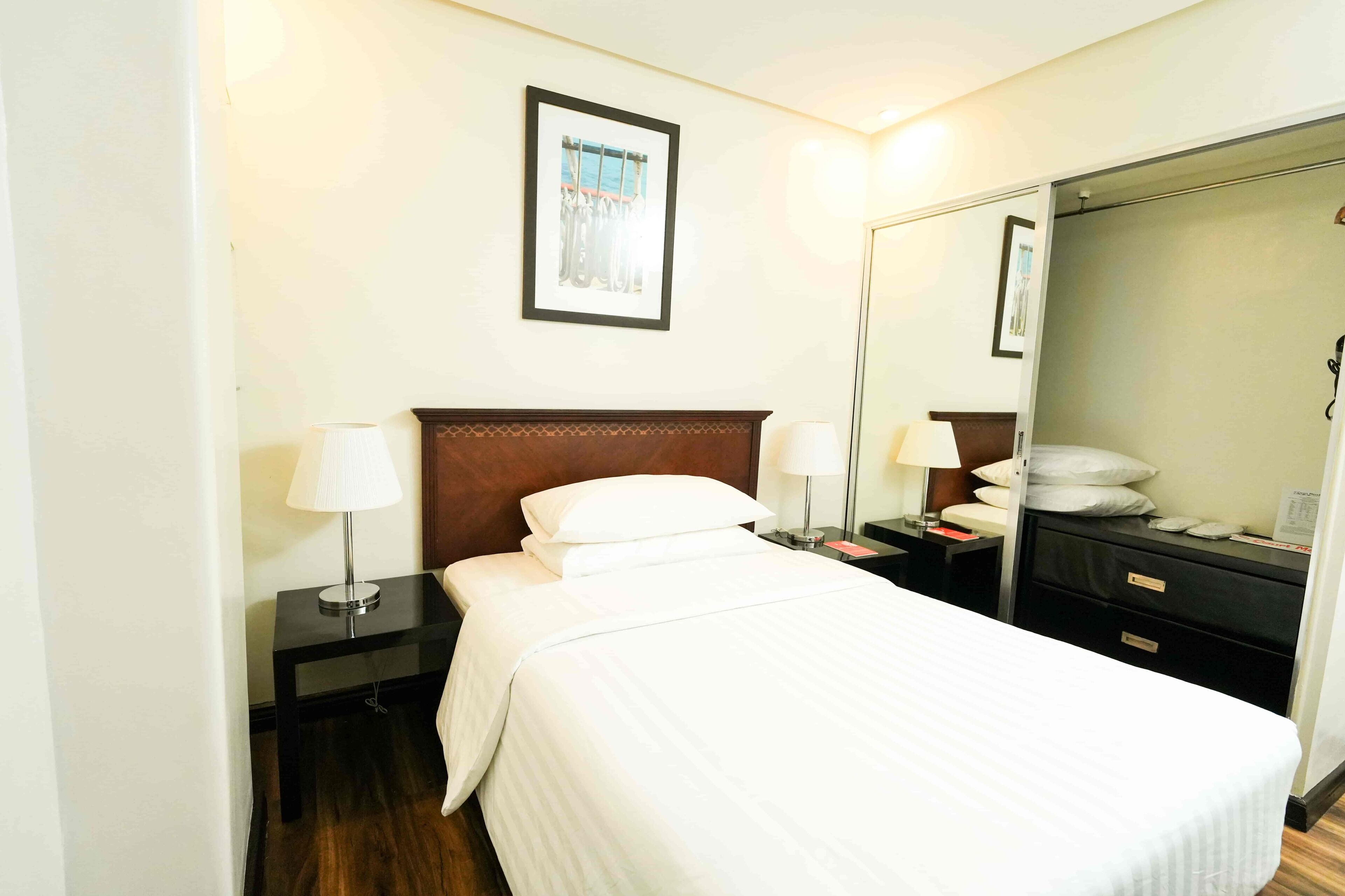 A neatly made bed with white linens, a wooden headboard, two bedside tables with lamps, a nightstand, a mirror, and a dresser with drawers.