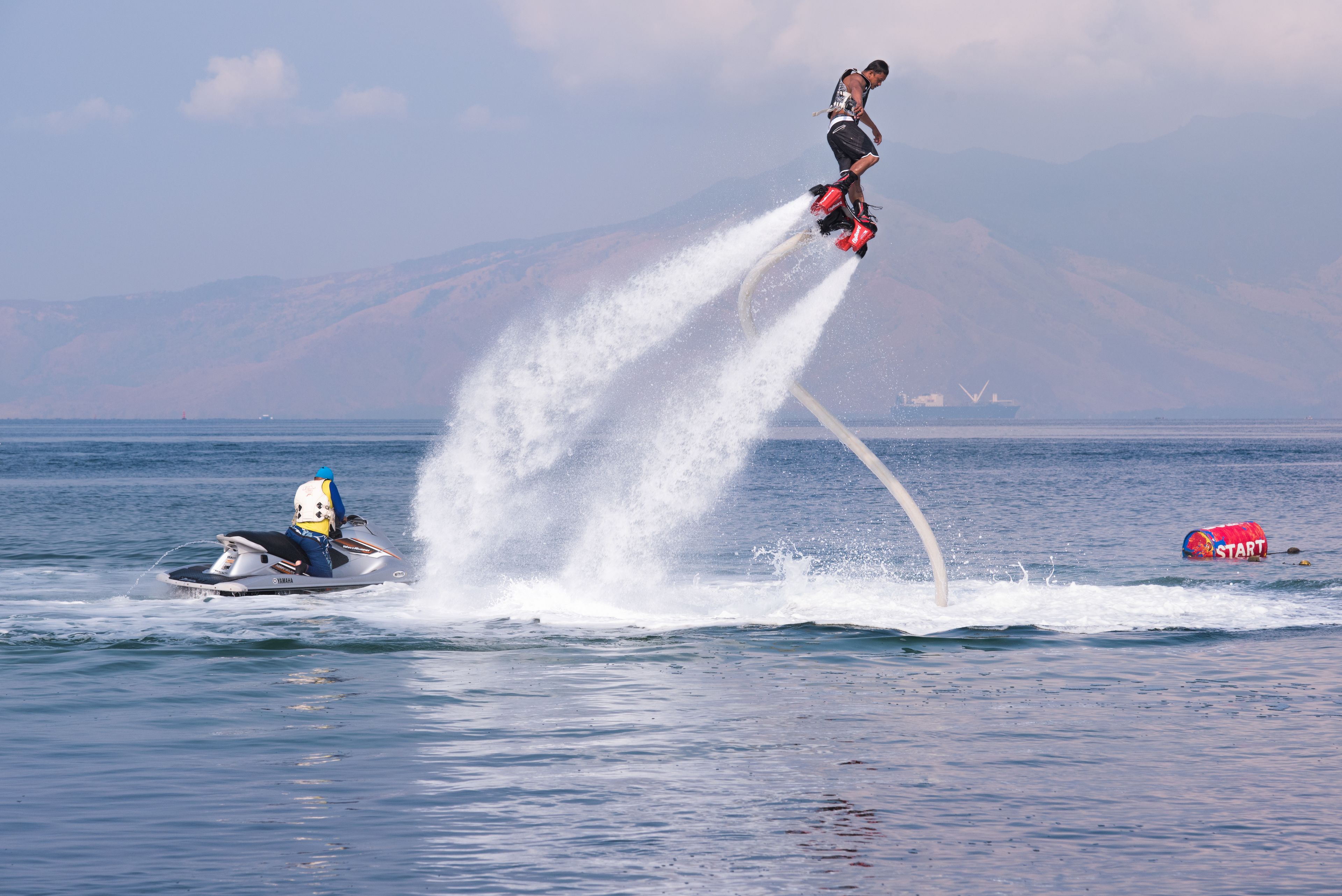 Un jet ski con una persona realizando un salto, salpicaduras de agua y un barco a lo lejos.