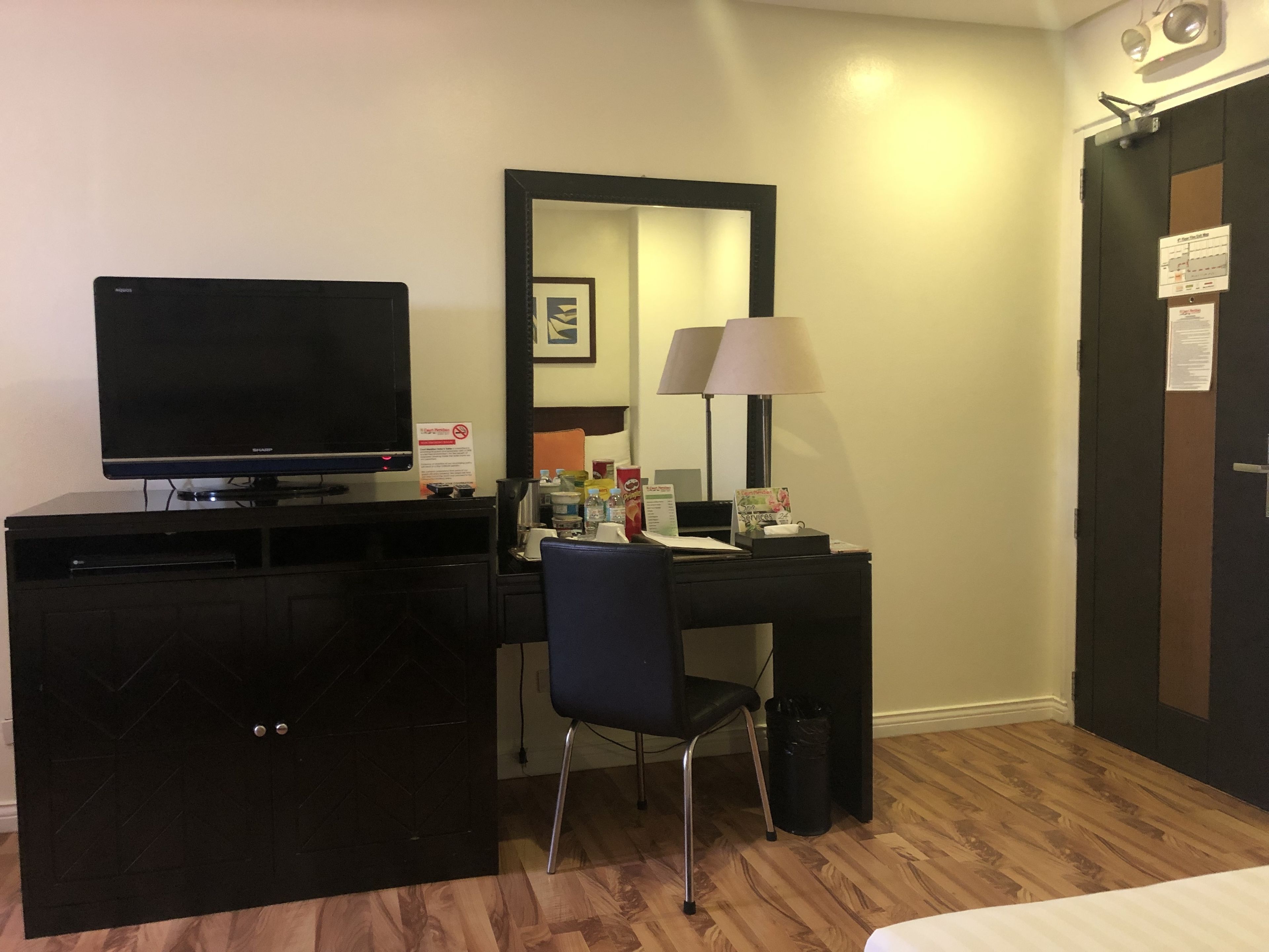 A hotel room with a flat-screen TV on a cabinet, a desk with a chair, a lamp, and a mirror.