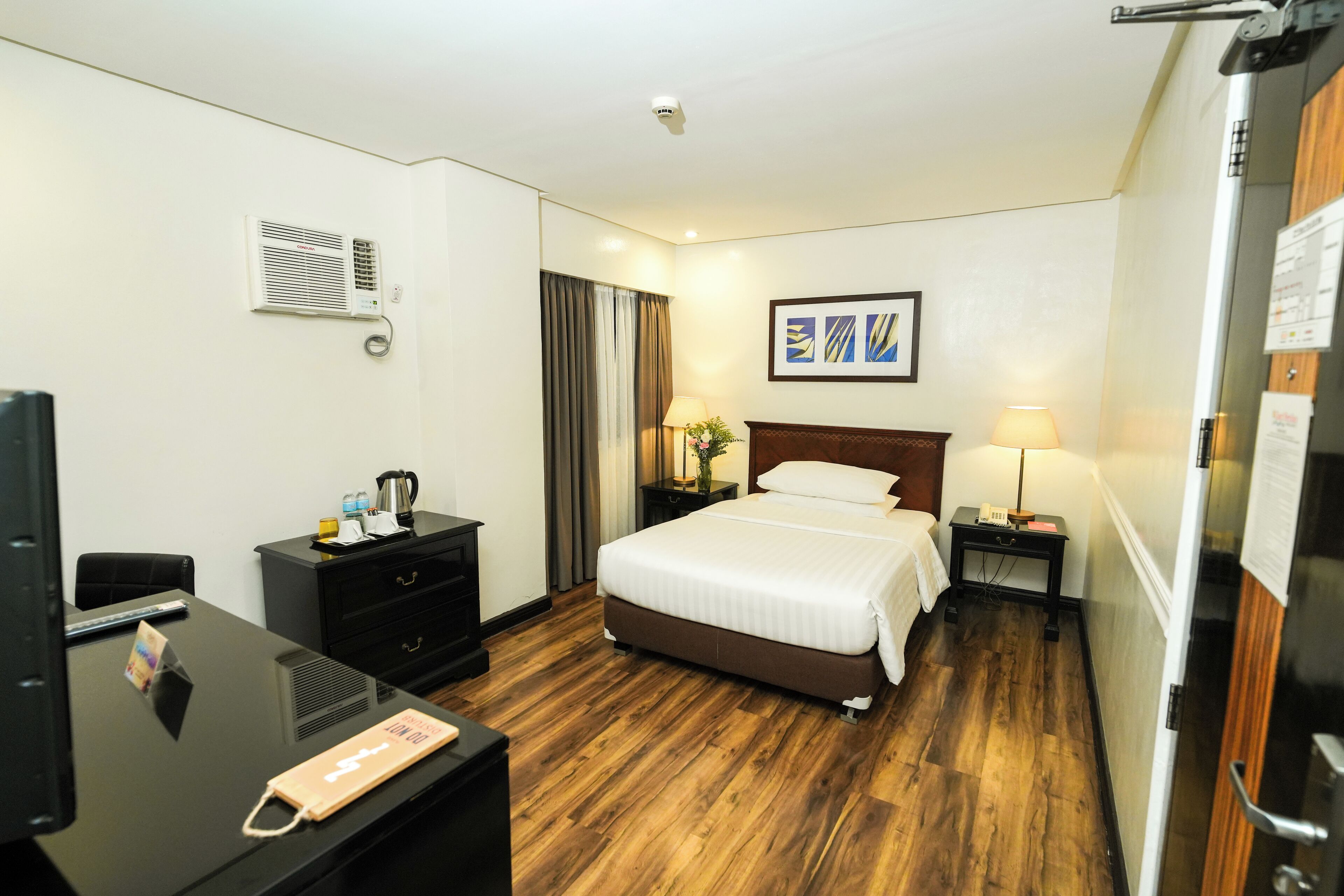Une chambre d’hôtel avec un lit, des tables de chevet, un bureau avec un ordinateur, une télévision et un plancher en bois.