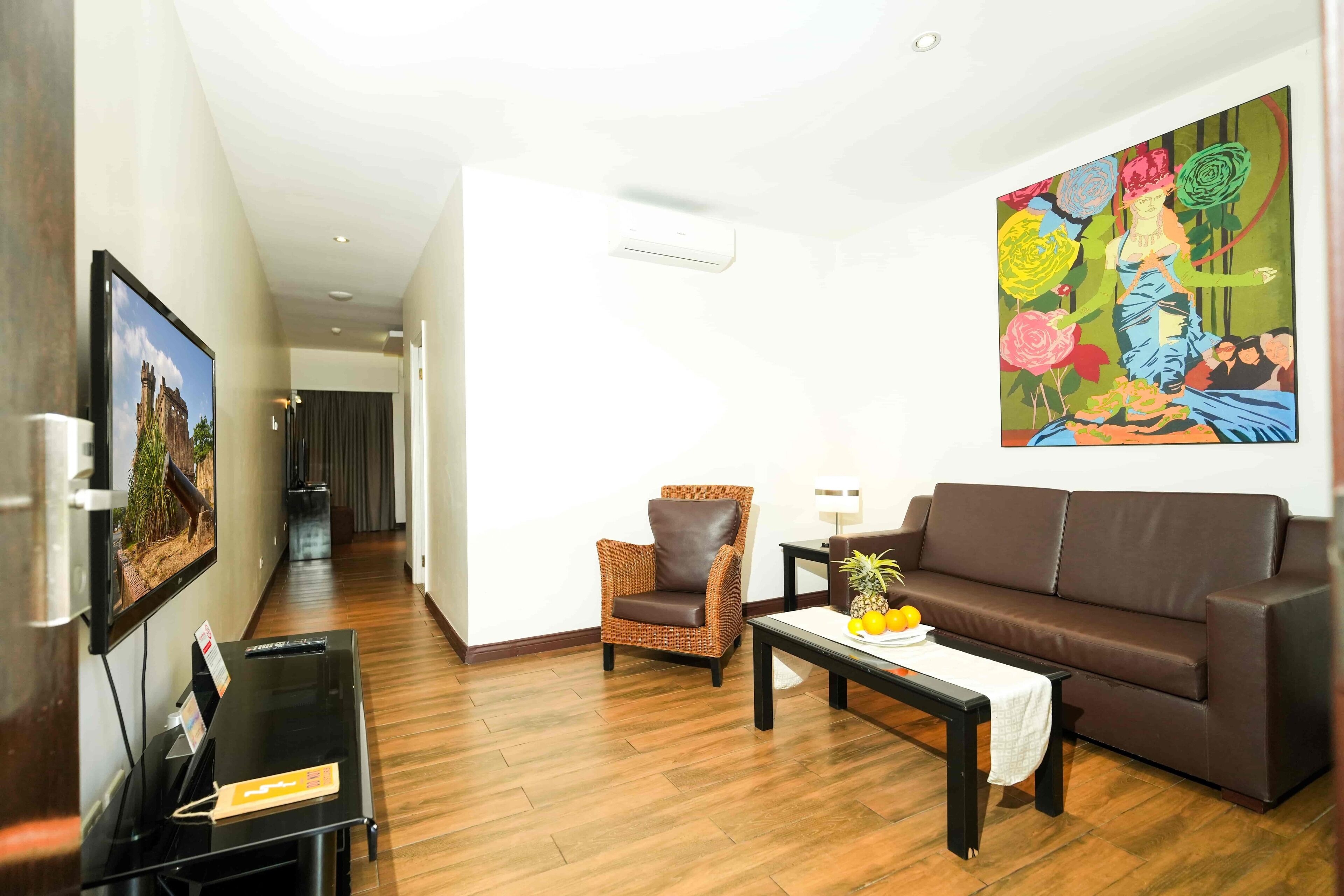 A living room with a brown leather sofa, a wicker armchair, a coffee table with fruits, and a painting on the wall.
