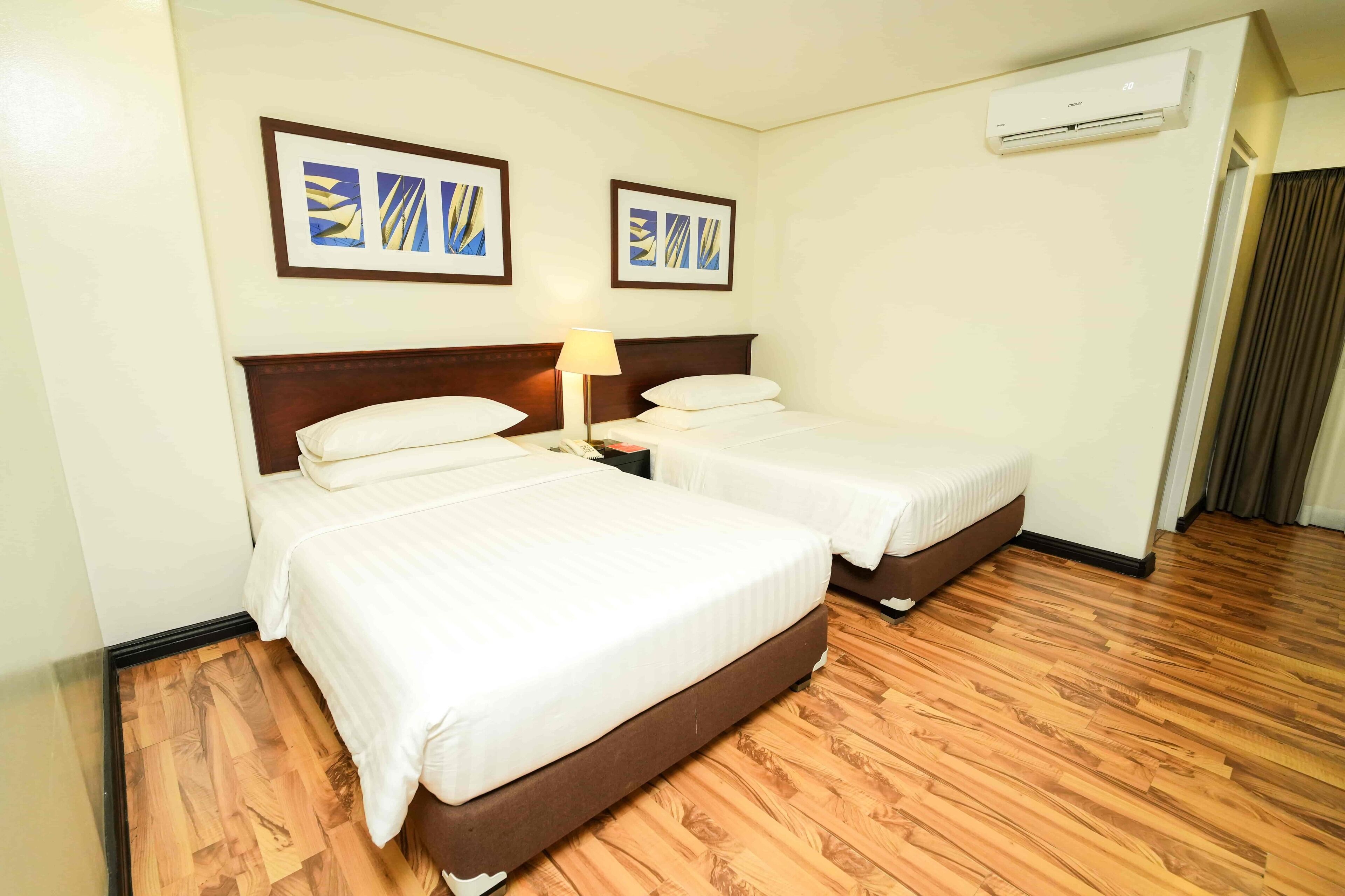 A hotel room with two beds, a wooden floor, and an air conditioning unit on the wall.