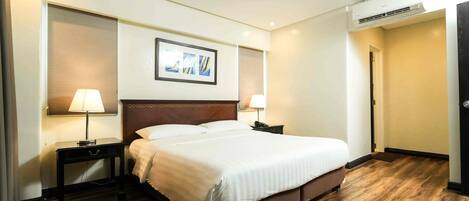 Habitación de hotel con piso de madera, una cama grande, dos mesitas de noche con lámparas, un cuadro enmarcado en la pared y un equipo de aire acondicionado.