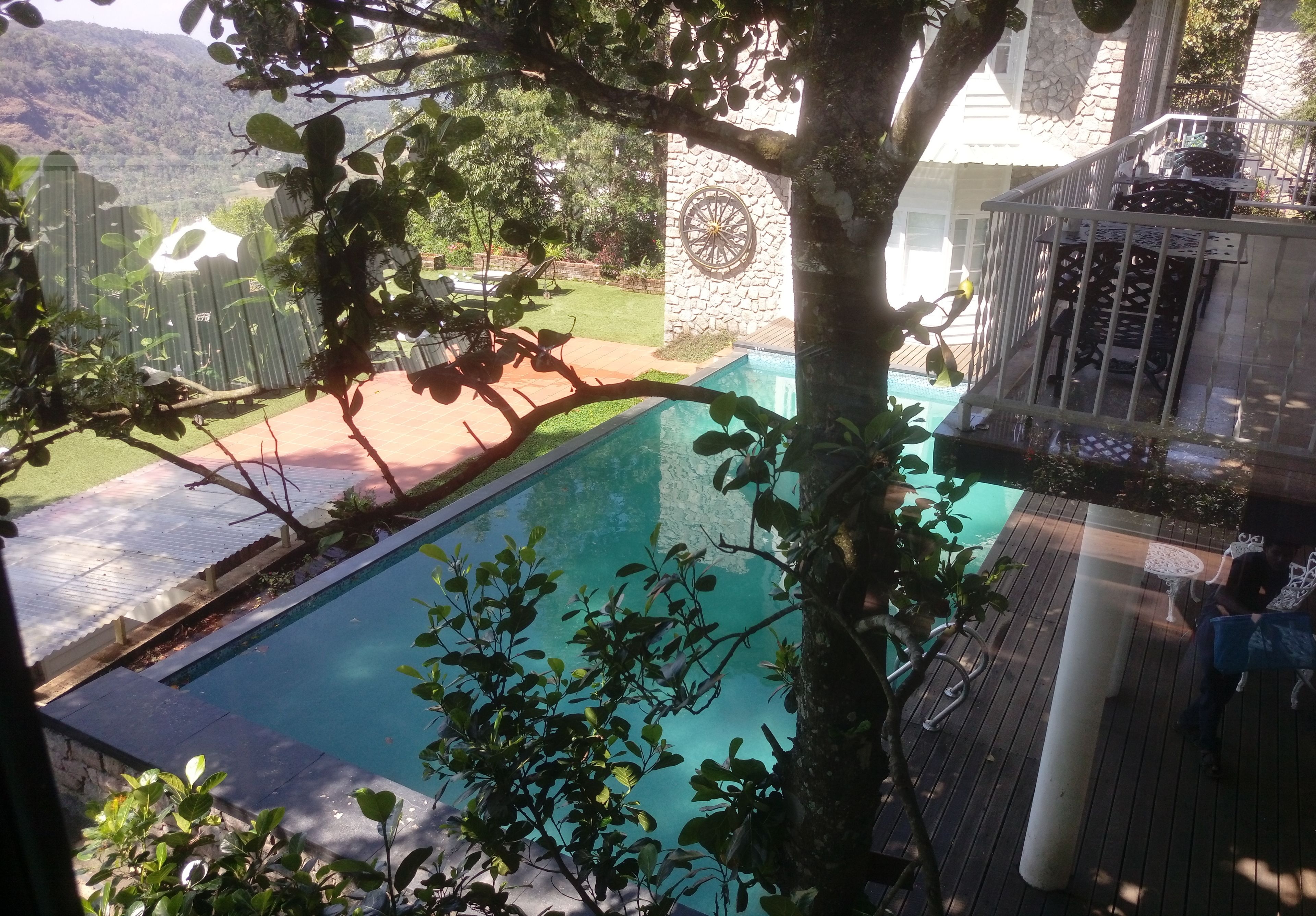 Una piscina rodeada de follaje, un muro de piedra con una rueda decorativa y una terraza de madera con mobiliario exterior.