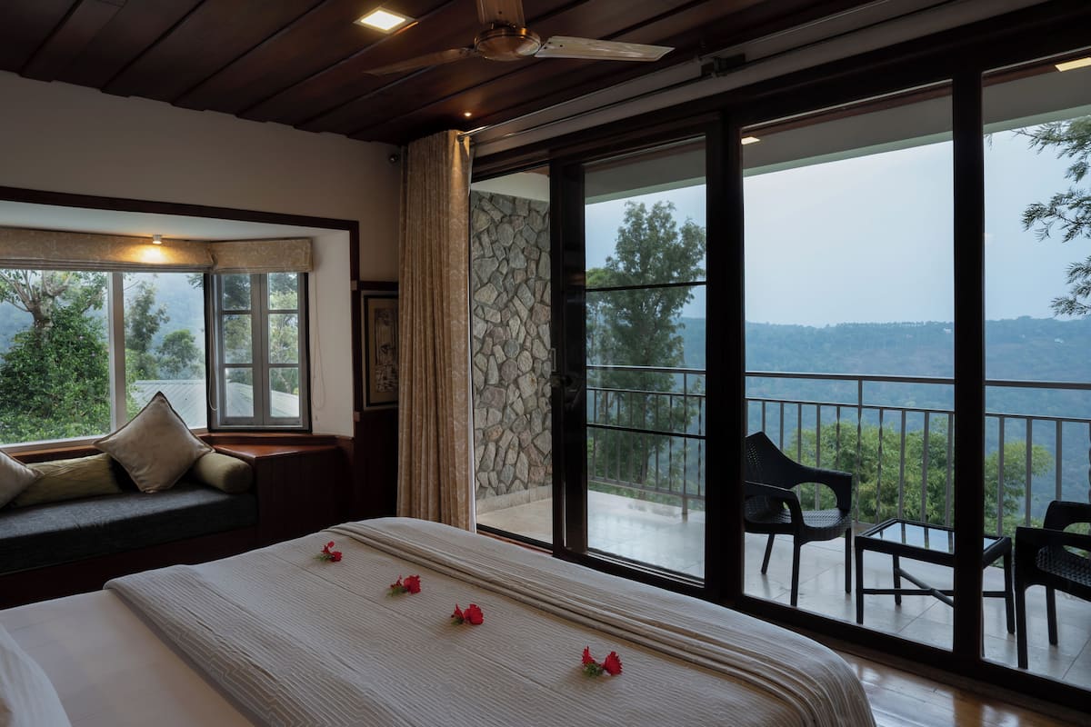 A bedroom with a large bed, a window with a view of trees, a stone wall, and a balcony with chairs.