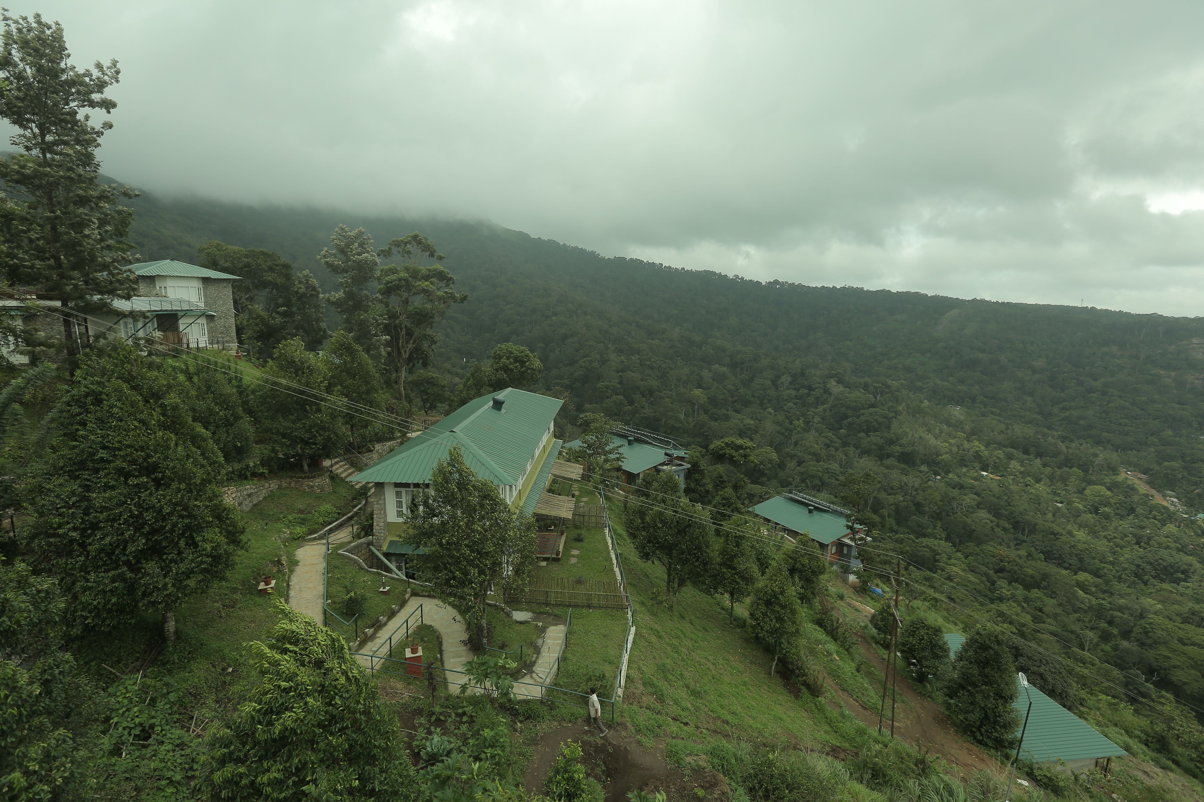 A hillside property with multiple buildings, including a main house with a green roof and a smaller structure with a similar roof. There are pathways and a fenced area visible.