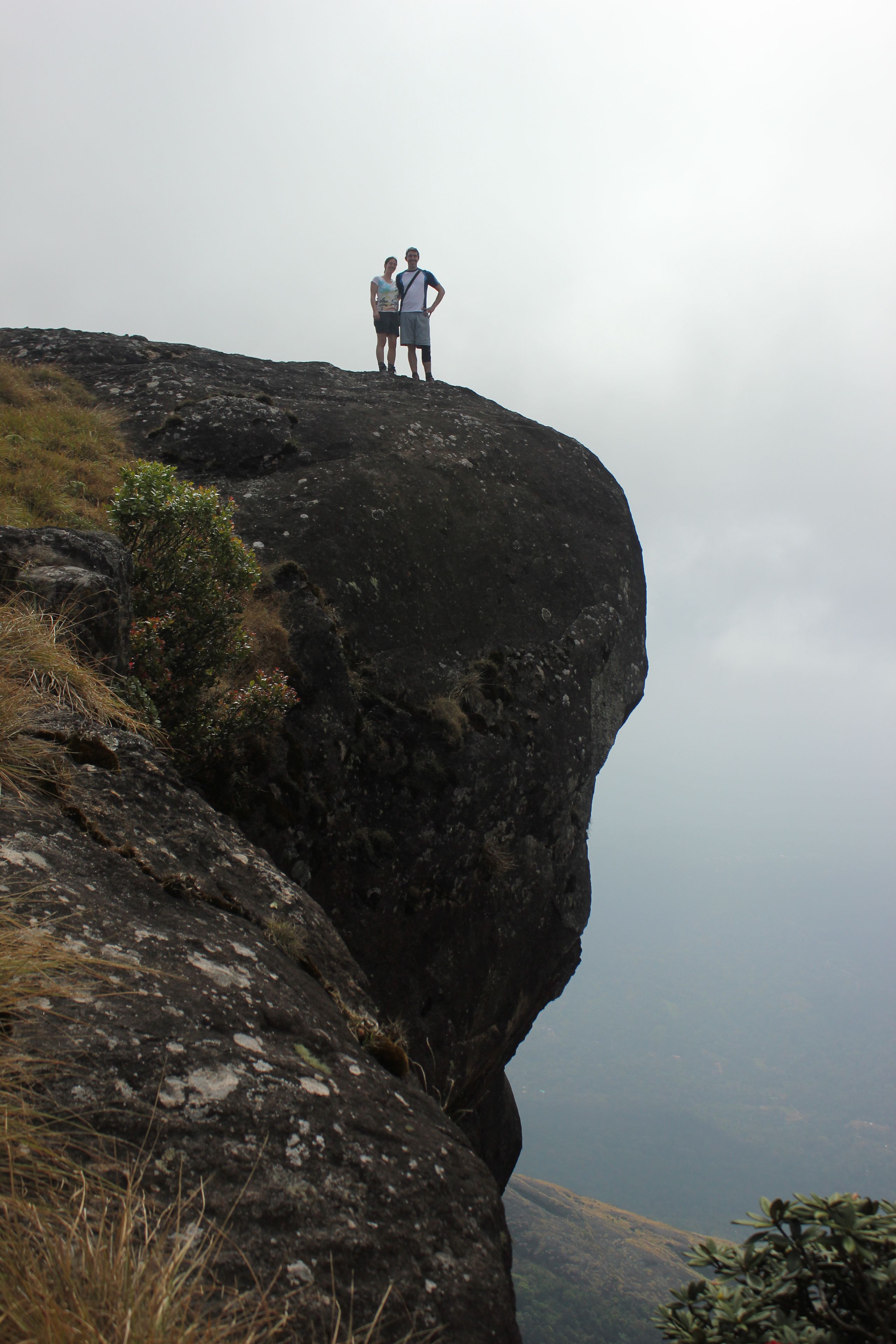 Dos personas de pie en el borde de un acantilado rocoso, con vistas a un valle.